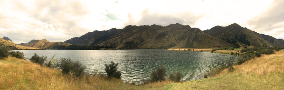 Panoramic view of Lake Moke near Queenstown New Zealand - Stunning mountain range landscape with crystal clear water and golden grassy shores under a dramatic cloudy sky