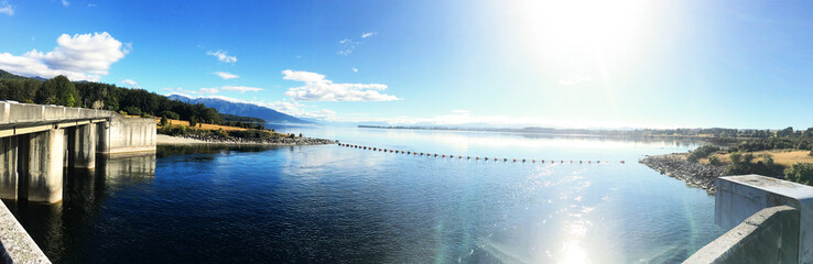 Obraz premium Stunning panoramic view of Lake Te Anau with Waiau River control gates - Scenic New Zealand landscape featuring bright morning sunlight, clear blue skies, and calm water