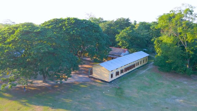 Drone view of school campus with long yellow building, open ground, and trees in Byaskuchi village, Assam.