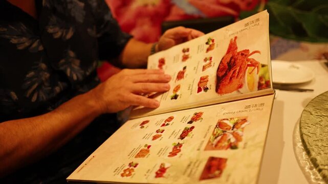 A male customer dressed in a stylish flowered shirt peruses a detailed menu at a refined Chinese restaurant with soft ambient lighting.