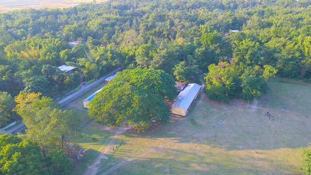 Drone view of school field with people, trees, nearby road, and buildings in Byaskuchi village, Assam.