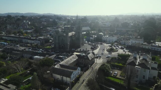 Monkstown Village, Ireland, revealing a Gothic church spire rising above historic buildings, coastal town rooftops, and rolling hills under bright sunlight.