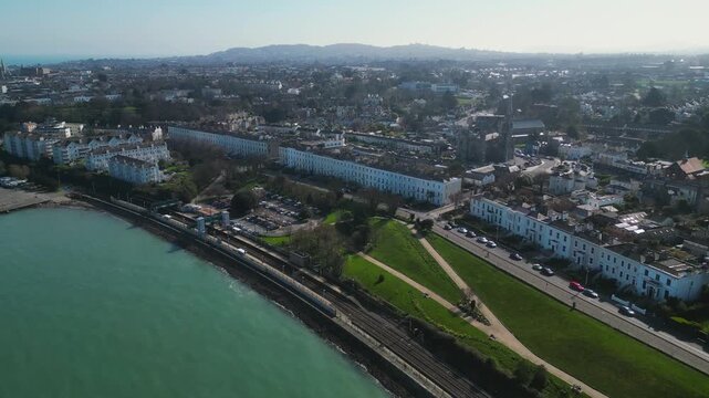 Aerial footage of Monkstown coastal suburb in Dublin, Ireland. Victorian terraced houses line the bay waterfront with urban cityscape, green parks, and mountain backdrop.