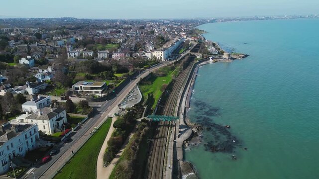 Drone footage of Monkstown coastal railway line along Dublin Bay. Train tracks run parallel to residential neighborhoods and turquoise ocean waters in Ireland.