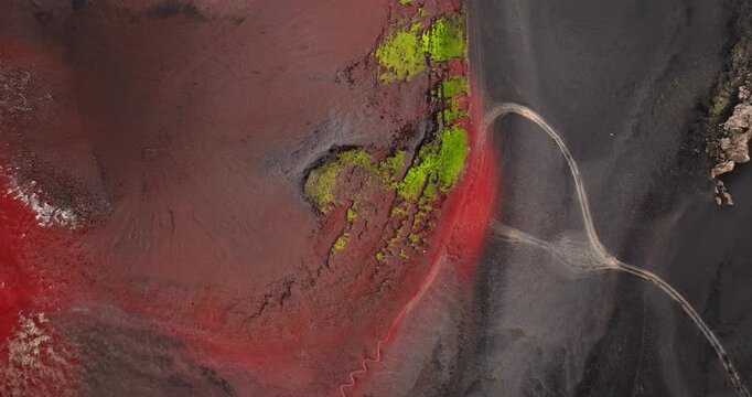 Top-down aerial of a red volcanic crater edge with a winding footpath cutting through black ash and bright moss, creating a striking abstract terrain pattern.