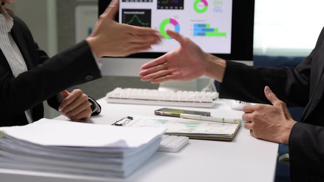 Business colleagues shaking hands after successful meeting in the office. Asian businessman presenting data on a computer to colleague, celebrating success with a handshake in office.