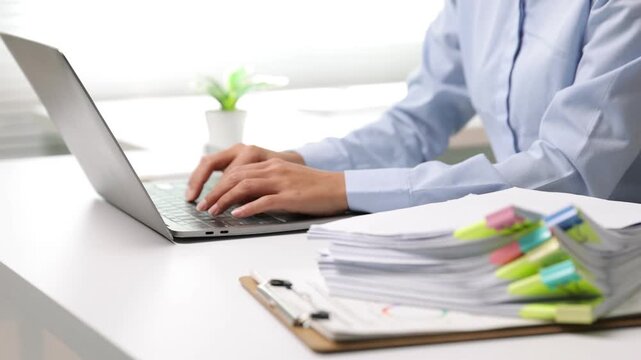 Office employee hands typing on laptop keyboard. Businesswoman working in the office, typing on a laptop with a stack of documents on the desk. Busy workflow concept.