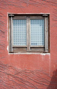 A weathered wooden double window set into a red stucco wall, framed by bare climbing vines and frosted glass with a square grid pattern, evoking rustic, textured urban architecture and winter