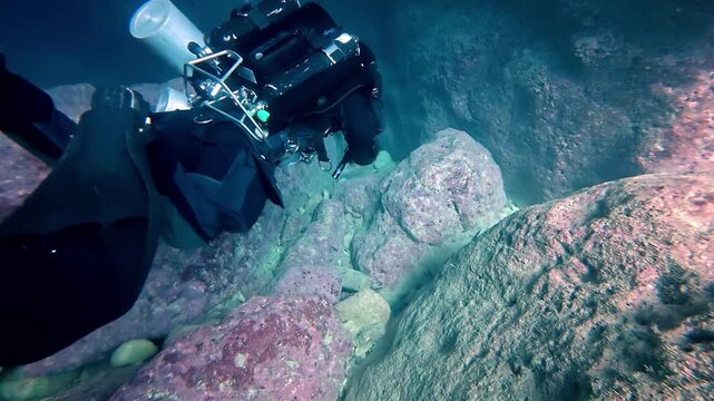 Professional technical scuba diver exploring deep underwater rocky cavern during summer season featuring dark blue marine tones and cinematic wide angle perspective with specialized rebreather gear