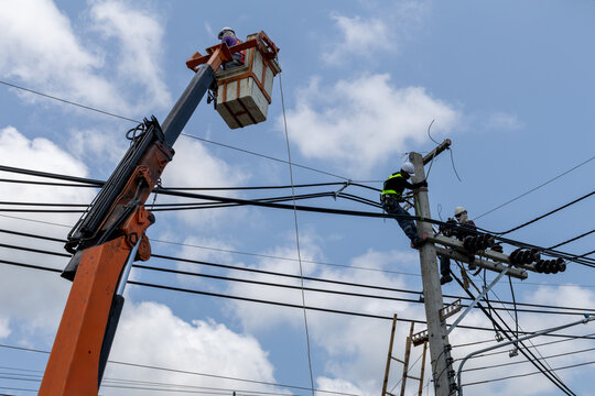 Utility workers repair and replace electrical wires on a tall pole in Thailand, using a bucket truck and safety gear to maintain a safe and operational power grid.