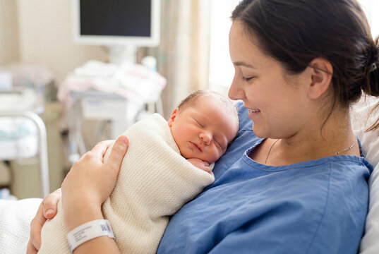 Mother holding her sleeping swaddled newborn baby on her chest in a hospital room