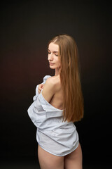 A young woman poses in a white off-the-shoulder shirt on a black background. The soft lighting highlights her relaxed and confident expression. She is wearing minimal underwear, which adds to her sens © SergeiPh