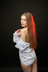 A young woman poses in a white off-the-shoulder shirt on a black background. The soft lighting highlights her relaxed and confident expression. She is wearing minimal underwear, which adds to her sens © SergeiPh