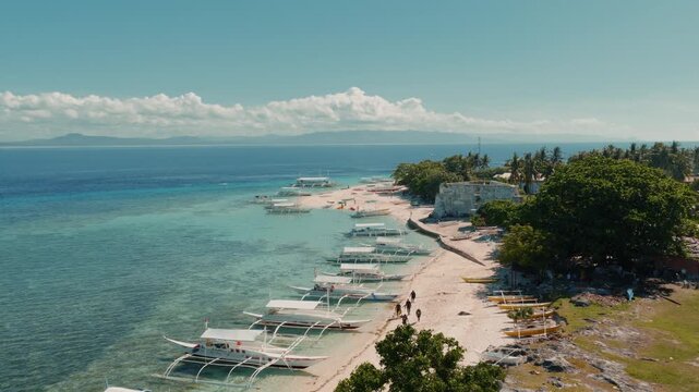 Tropical beach coastline with rows of traditional outrigger boats anchored in clear shallow sea, aerial dolly along shoreline