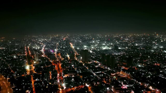 An elevated, wide-angle view of a massive, illuminated city skyline at night. Multiple colorful fireworks displays explode across the dense urban sprawl, celebrating New Year's Eve