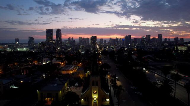 Drone flying backward from the top of a church revealing the San Diego skyline at 8PM during sunset with warm evening tones.