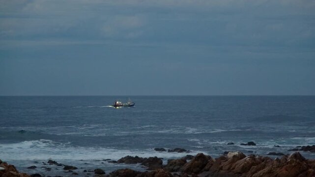 Fishing vessel with lights on at twilight cruise out on ocean near rocky shore