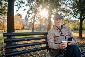Fototapeta premium Senior man relaxing on park bench enjoying coffee and autumn day