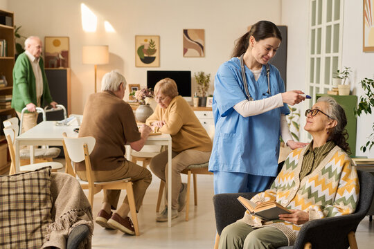 Senior woman receiving temperature check from female nurse while reading in care home, with elderly residents socializing behind. Ideal for eldercare, assisted living, healthcare support marketing