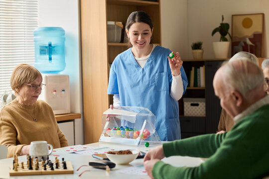 Young adult female caregiver leading bingo activity with seniors in care center, supporting cognitive engagement and social interaction. Useful for elder care, memory support, retirement services