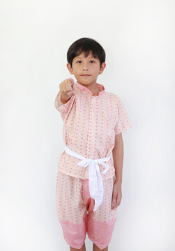 A young Thai boy, wearing a traditional pink costume with a white sash, is standing against a white background and pointing his finger forward with a confident expression.