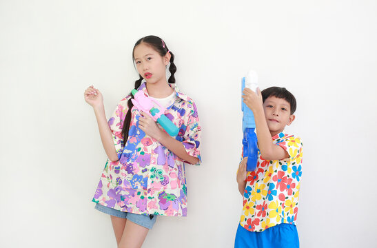 Two children, a girl and a boy wearing colorful floral shirts, are having fun playing with water guns against white background, Ready for Songkran festival celebrations.