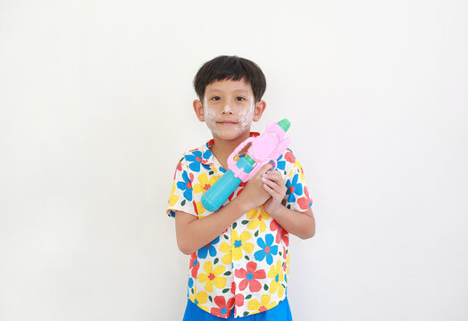 A young Thai boy, with face powder for the Songkran festival, is smiling and holding colorful water gun against white background.