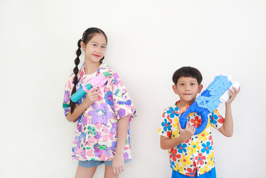 Two children, a girl and a boy wearing colorful floral shirts, are having fun playing with water guns against white background, Ready for Songkran festival celebrations.