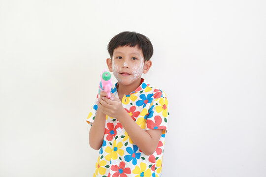 A young Thai boy, with face powder for the Songkran festival, is smiling and holding colorful water gun against white background.