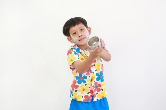 Thai boy, with white facial powder applied for Songkran festival and wearing colorful floral shirt, is standing and holding a small silver bowl, getting ready to splash water.