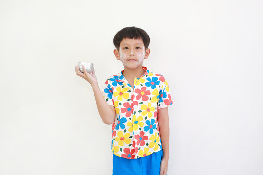Thai boy, with white facial powder applied for Songkran festival and wearing colorful floral shirt, is standing and holding a small silver bowl, getting ready to splash water.