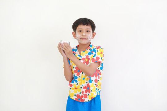 Thai boy, with white facial powder applied for Songkran festival and wearing colorful floral shirt, is standing and holding a small silver bowl, getting ready to splash water.