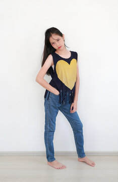 Confident young Asian girl posing in stylish navy blue fringe tank top with a yellow heart and casual denim jeans against white wall.