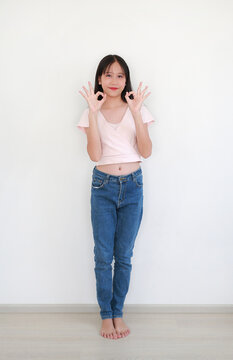 Cheerful young Asian girl, wearing light pink wrap-style crop top and blue denim jeans, standing barefoot and making 'OK' hand gestures with both hands while smiling on white wall.