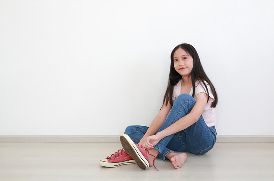 Portrait of young Asian girl with long dark hair in pink t-shirt and blue jeans, sitting on the floor while putting on her red sneakers against clean white wall.