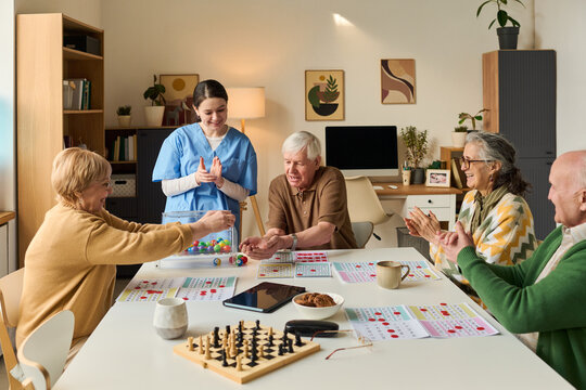 Senior group playing bingo with female caregiver, interacting around activity table in care facility. Useful for elder care, assisted living, social support, retirement services marketing