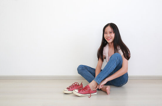 Portrait of young Asian girl with long dark hair in pink t-shirt and blue jeans, sitting on the floor while putting on her red sneakers against clean white wall.