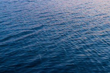 Obraz premium Blue Water Surface with Gentle Ripples and Sunset Reflections,A high-angle, full-frame shot of deep blue water featuring rhythmic ripples and soft pinkish-golden reflections from the evening sun,