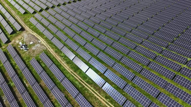 Rows of solar panels stretch across a green field. Sunlight reflects off the panels. The sky is clear with no clouds. This area is situated in a remote location.