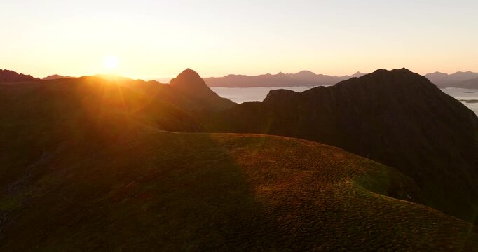 Golden sunrise light illuminating Storheia mountain ridge above coastal landscape in Norway, aerial ascend overview