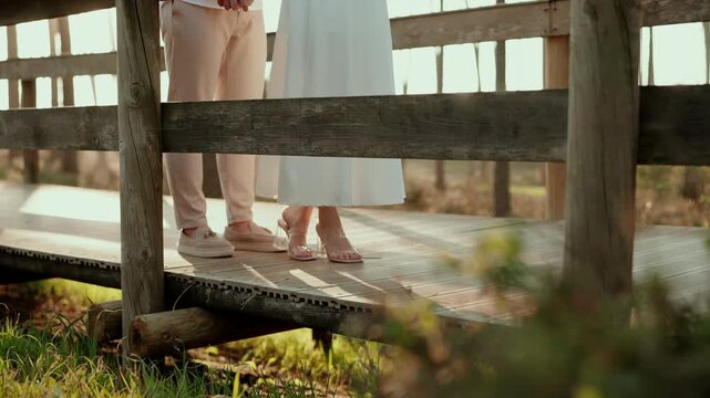 Low angle shot through rustic wooden fence of bride in clear heels and white gown beside groom in loafers standing on a sunlit wooden boardwalk