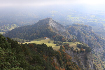Fototapeta premium Misty mountains roll beneath a hazy, sunlit sky. Deep green pines frame rugged, rocky peaks. Autumn hues paint slopes in gold and russet. Lone cottage sits in a tranquil valley clearing