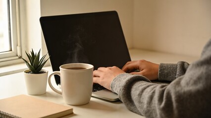 Fototapeta premium Minimalist desk setup featuring a laptop, a steaming cup of coffee, and a small indoor plant near a window. Represents productivity, calm lifestyle, and modern work environment.