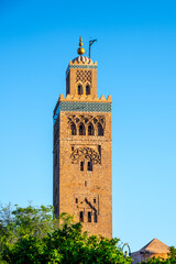 Naklejka premium The tower of Kutubiyya Mosque stands tall in Marrakesh, Morocco. The clear blue sky forms a backdrop as the sun shines on this historical site. Visitors may be nearby, enjoying the scene.
