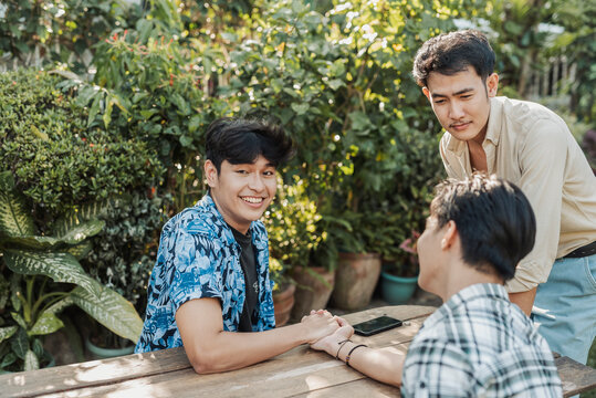 A playful young Asian man smiling proudly after winning a friendly arm wrestling match against his friend during a fun outdoor hangout. Outdoor shot.