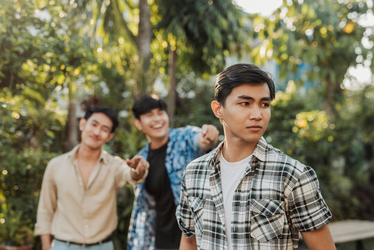 A annoyed young Asian man ignoring two friends pointing and laughing at him during a teasing and mocking moment. Outdoor shot.
