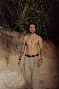 Shirtless tattooed man smiling at camera by pandanus on beach
