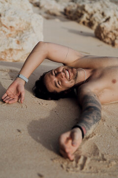 Tattooed man lying on sand smiling at camera with arm outstretched