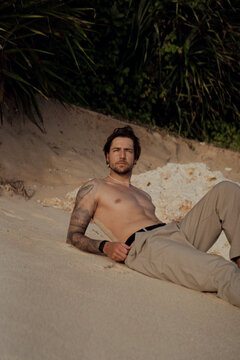 Shirtless tattooed man sitting on sand leaning back at beach cliff