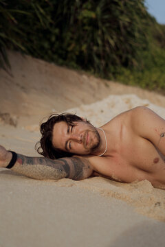 Shirtless tattooed man lying on side on sand smiling at camera